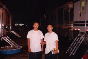 Two men in white t-shirts standing outside production trailers at night, taking a break from filming taekwondo sequences and intense reelstunt performances.